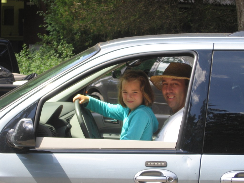 A young girl sitting on her father's lap in the front seat of the family car pretending to steer it